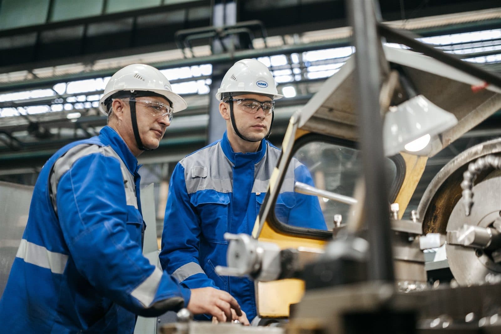 Dois operários com capacete e óculos de proteção examinam juntos uma máquina industrial.